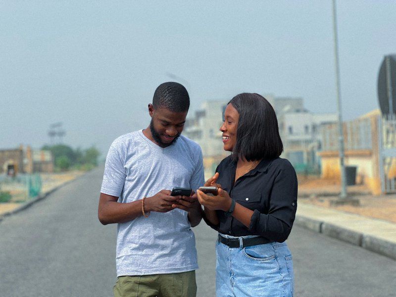 A young man and woman standing on a street in Lagos, smiling as they look at their phones together.